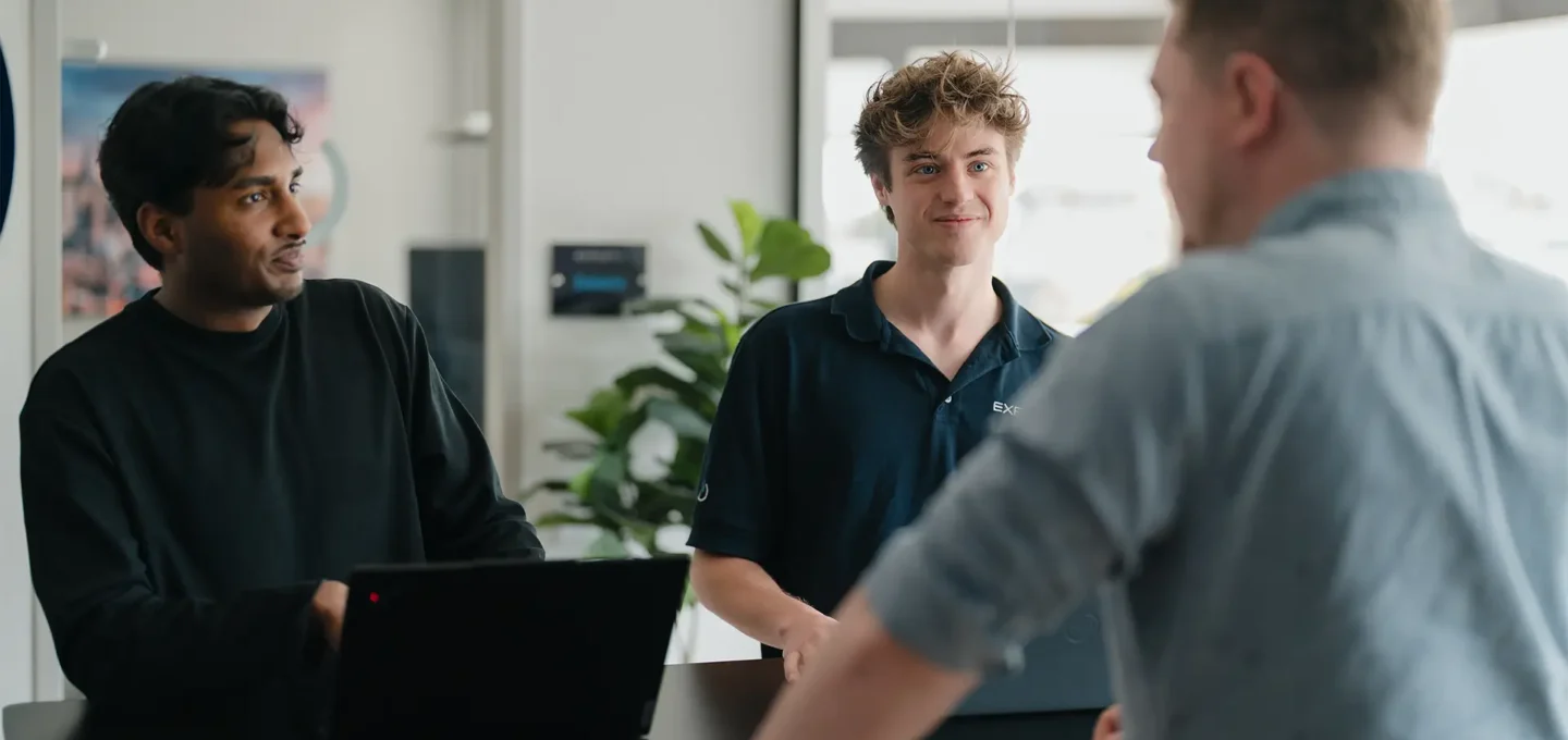Drei EXPLETO-Mitarbeiter tauschen sich gemeinsam an einem Arbeitsplatz mit Laptops aus und besprechen ein Projekt in einer modernen Büroumgebung.