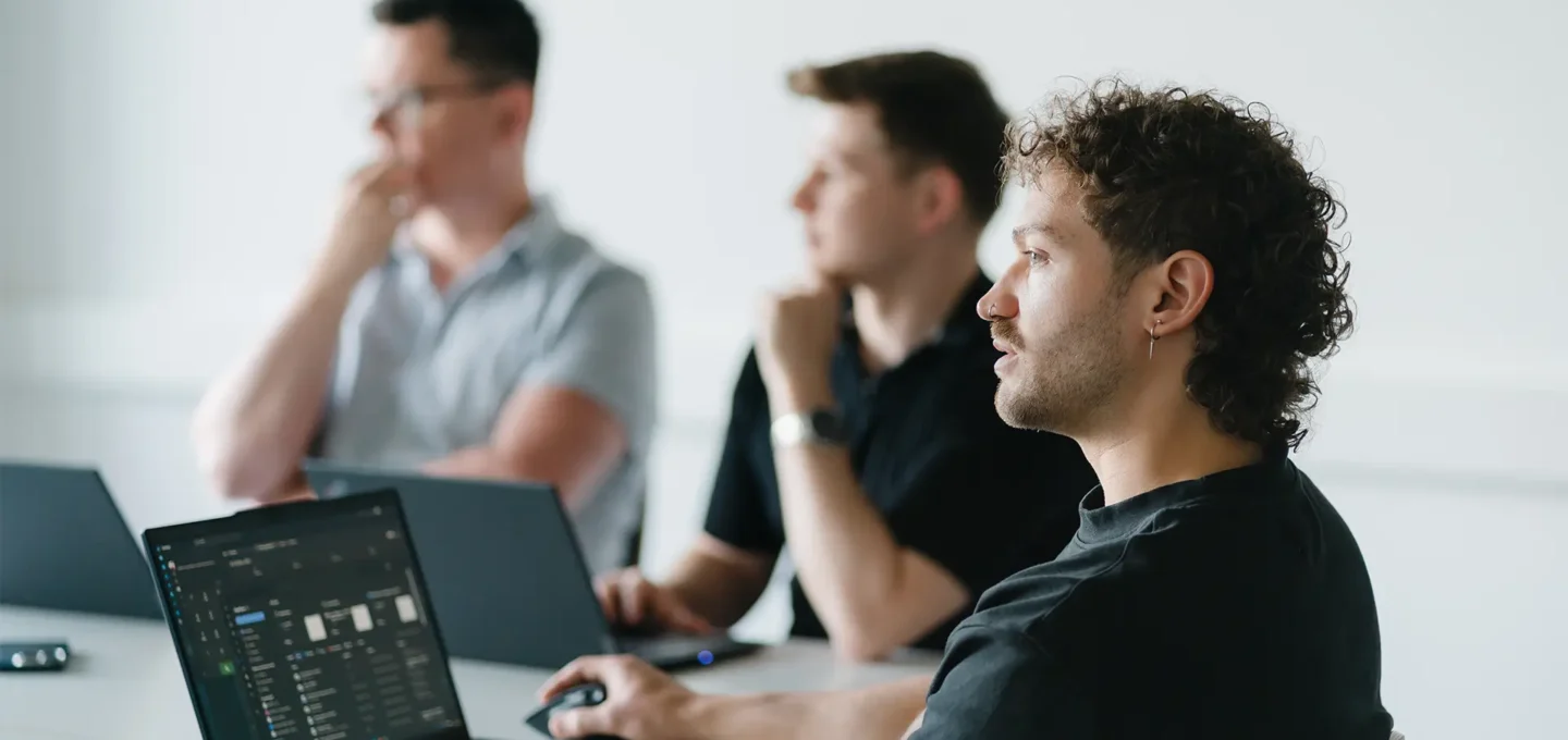 Drei EXPLETO-Mitarbeiter arbeiten konzentriert an ihren Laptops und tauschen sich während einer gemeinsamen Besprechung in einer modernen Büroumgebung aus.